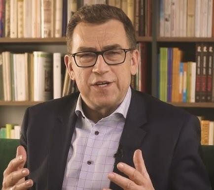 Maciej Orłoś z jedną ze swoich książek w domowym gabinecie, otoczony półkami z książkami.