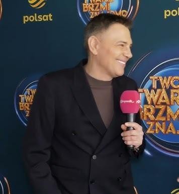 Krzysztof Ibisz prowadzi dynamiczny program telewizyjny w studiu pełnym świateł i ekranów.